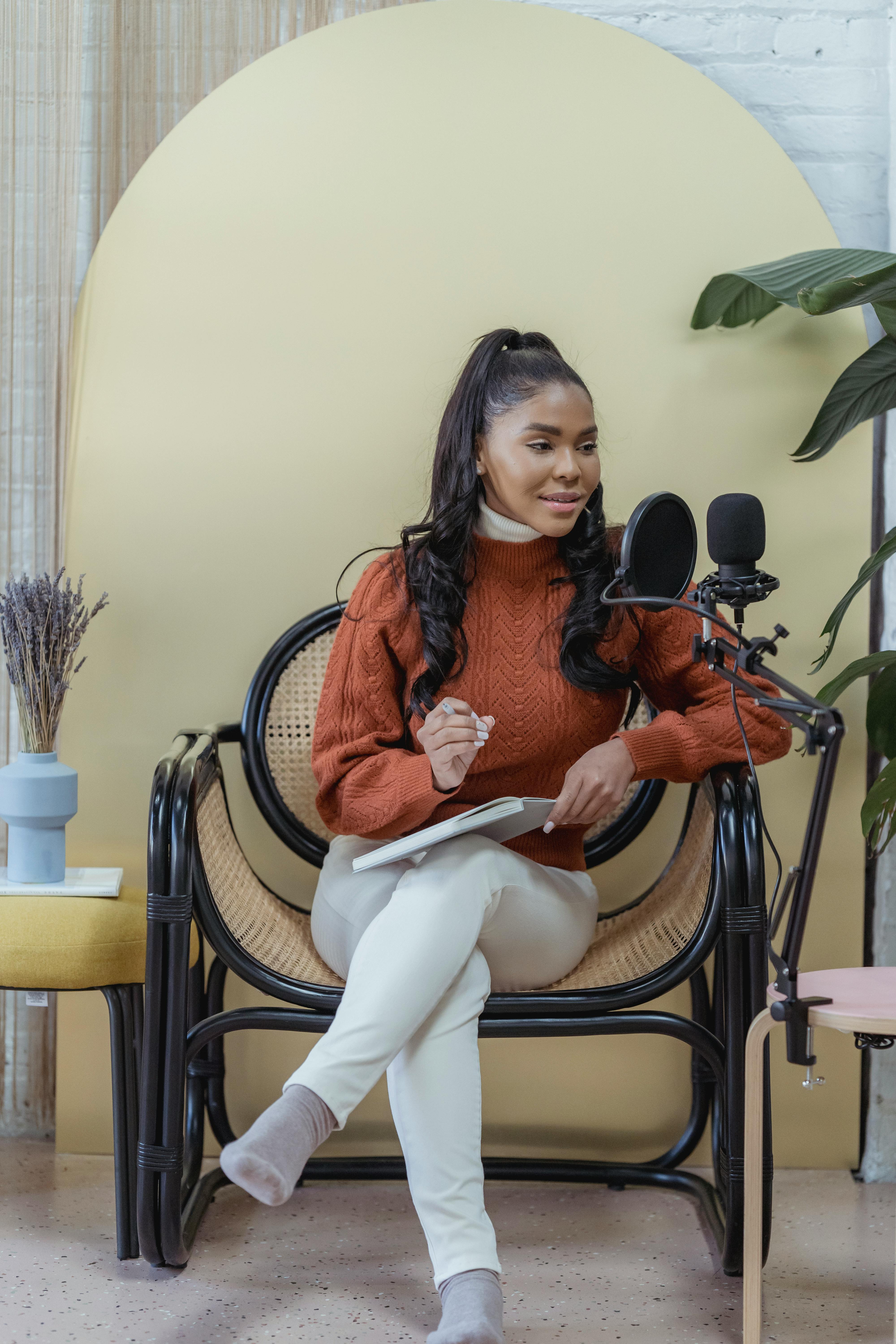 A young woman sits in a stylish home studio, recording a podcast with a microphone.